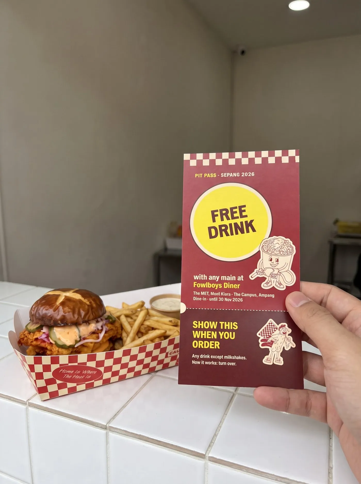 A red pit pass voucher for a free drink at Fowlboys Diner, held over a sandwich and fries